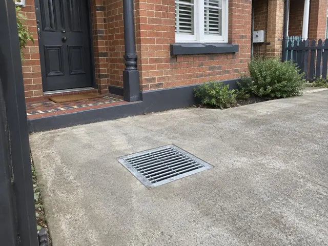 Outdoor drain grate set in dry concrete paving beside a brick house in Botany